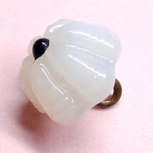 May include: A translucent white, pumpkin-shaped button with a glossy finish and vertical ridges. The button features a small, dark black circular accent at the top center and a metallic bronze-toned shank loop extending from the bottom side.