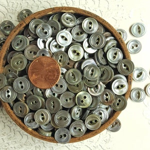 May include: A bowl filled with a large quantity of iridescent, two-hole, round buttons. The buttons are a light green color with a silver sheen. A penny is included for scale.