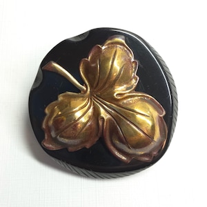 May include: A round, black button featuring a raised, metallic gold-toned three-lobed leaf design in the center. The button has a smooth, dark surface with subtle, diagonal etched lines along the outer edge, providing a textured border.