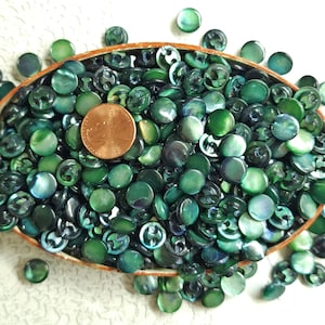 May include: A bowl filled with green iridescent shell buttons. The buttons are round and have a slight sheen. There is a US penny in the bowl for scale.