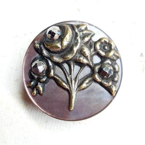 May include: A round, antique button with a floral design. The button features a raised bronze-colored bouquet of roses and other flowers, with faceted crystal accents. The button has a dark mauve background and a bronze rim.
