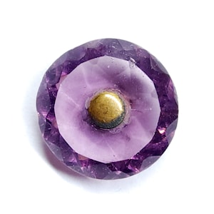 May include: A circular button featuring a translucent purple faceted glass outer ring and a smooth, solid brass-colored metal center. The design incorporates a beveled edge with multiple geometric facets that catch light, surrounding the central metallic dome.