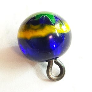 May include: A spherical glass button featuring a smooth, polished surface with swirling bands of deep blue, bright yellow, and vibrant green colors. The button includes a single metal shank loop attached to the bottom for secure fastening.