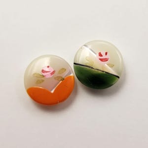 May include: Two round, vintage buttons. One button has a pink rose design with orange accents, the other has a pink rose design with green accents. Both buttons are translucent with a white background.