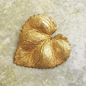 May include: A gold-toned brooch shaped like a heart-shaped leaf. The surface features a detailed, raised texture that mimics the natural veins and organic contours of a real leaf, with a polished metallic finish that reflects light across its surface.