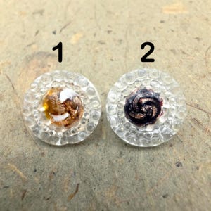 May include: Two round buttons with a clear, textured outer rim featuring small raised circles. Button 1 has a center with amber, white, and copper-colored swirls. Button 2 has a center with dark purple and copper-colored swirls. Both are labeled 1 and 2.