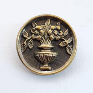 May include: A round, antique-style button with a raised floral design. The button features a central vase overflowing with flowers and leaves, set against a dark background with a gold-colored rim. The button is made of metal.