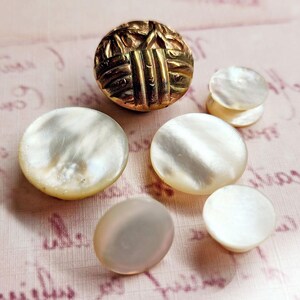 May include: A collection of six buttons, five are white mother of pearl and one is gold with a geometric design.
