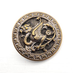 May include: A round, antique-style button with a detailed design. The button features a central image of a griffin with outstretched wings, in shades of gold and bronze. The griffin is surrounded by an ornate border. The button is likely made of metal.