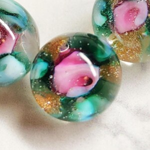 May include: Close-up of three glass beads with floral designs. Each bead features a pink rose encased in clear glass, surrounded by green, gold, and teal accents. The beads have a rounded shape and a glossy finish, creating a unique and artistic look.