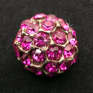 May include: A spherical bead covered in numerous round, faceted hot pink crystals set into a metallic gold-toned base. The surface of the bead features a dense, honeycomb-like arrangement of these sparkling, vibrant pink stones across its entire exterior.