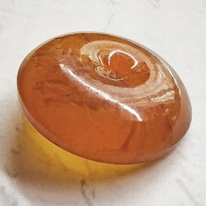 May include: A smooth, oval-shaped amber-colored stone with a glossy finish. The stone has a swirl pattern on the top surface and casts a yellow shadow. The object is likely a decorative item or a piece of jewelry.