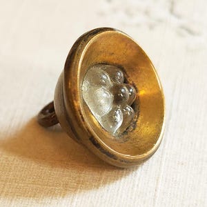 May include: A vintage brass bell with a clear, textured glass insert. The bell has a rounded shape with a small loop for hanging. The glass insert has a cluster of bubble-like shapes.
