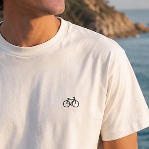 Op de afbeelding: Een off-white T-shirt met korte mouwen en een ronde hals, voorzien van een klein, zwart geborduurd icoon van een fiets op de linkerborst. Het kledingstuk heeft een eenvoudig en strak ontwerp met een standaard ronde halslijn.