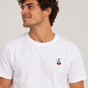 Könnte beinhalten: Ein weißes T-Shirt mit kurzen Ärmeln und einer kleinen Stickerei auf der linken Brust. Das Motiv zeigt eine rote Seilbahngondel an einem schwarzen Seil und präsentiert ein schlichtes Design auf einem einfarbigen, weißen Baumwollstoff.