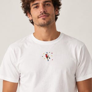 Könnte beinhalten: Ein weißes T-Shirt mit kurzen Ärmeln und einer kleinen, mittigen Stickerei, die eine Person beim Klettern an einer Wand mit bunten Griffen zeigt. Das Motiv stellt einen Kletterer in rotem Shirt und blauer Hose an verschiedenen Klettergriffen dar.