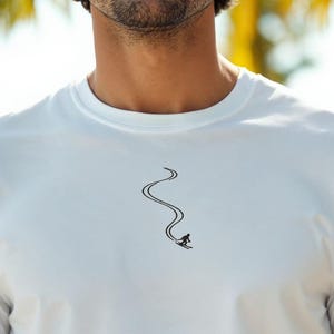 Op de afbeelding: Een wit T-shirt met korte mouwen en een kleine, centraal geplaatste zwarte afbeelding van een skiër die een bochtige lijn door de sneeuw trekt. Het minimalistische ontwerp bevindt zich op de borst en toont een skiër in beweging.
