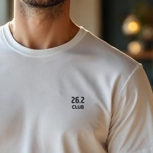 Pode incluir: Uma camiseta branca de gola redonda com o texto "26.2 CLUB" impresso em preto. A camisa é feita de um tecido macio e claro e está sendo usada por uma pessoa.