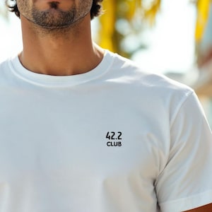 Könnte beinhalten: Weißes T-Shirt mit Rundhalsausschnitt und dem Text "42.2 CLUB" in Schwarz aufgedruckt. Das Shirt besteht aus einem weichen, leichten Material und hat kurze Ärmel. Das T-Shirt ist ein Freizeitartikel.
