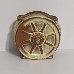 Puede incluir: Un jarrón de cerámica con diseño de rueda. El jarrón es de color beige claro con detalles marrones, y el diseño de la rueda se muestra prominentemente en la parte delantera. El jarrón tiene una base cuadrada y una parte superior redondeada, aprox. 10 cm de alto.