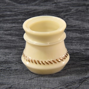 Puede incluir: Un pequeño recipiente de cerámica blanco con un borde dorado alrededor de la parte superior e inferior. El recipiente tiene una forma redonda y lisa y es perfecto para guardar objetos pequeños.