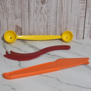 Puede incluir: Se muestra una cuchara medidora amarilla de doble extremo, un cuchillo rojo con sierra y una herramienta naranja con forma de cuchillo. Las herramientas son de plástico y están diseñadas para uso en la cocina. El fondo es una superficie de mármol blanco.