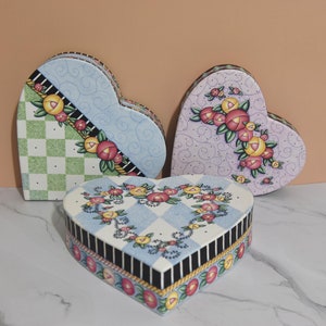 Puede incluir: Tres cajas en forma de corazón con diseños florales. Las cajas están hechas de cartón y tienen un acabado pintado. Las cajas son de diferentes colores y patrones. Las cajas están sobre una superficie de mármol blanco.