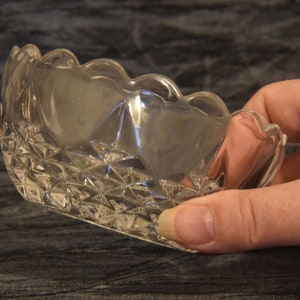 May include: A clear, scalloped-edge glass dish with a diamond-patterned base. The dish is held in a hand, showcasing its delicate design and transparency. The dish is likely for serving small portions.