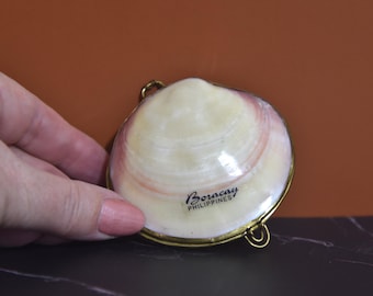 May include: A small, white seashell with a pink rim and a brass clasp. The shell has the words "Boracay Philippines" printed on it.