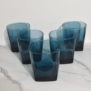 May include: Five teal-colored drinking glasses. Each glass has a square base and a slightly tapered, cylindrical shape. The glasses are arranged in a group, showcasing their design and color.