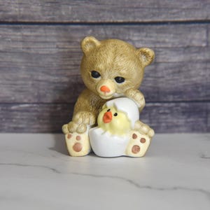May include: A ceramic figurine of a tan teddy bear holding a yellow chick hatching from a white egg. The bear has a black nose and eyes, and the chick has an orange beak. The figurine is on a white surface.