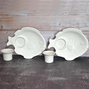 May include: Two white ceramic fish-shaped plates with a circular indentation and two small matching cups. The plates have a textured design and are set against a wooden background.