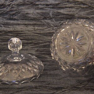 May include: A clear, cut-glass candy dish with a lid. The lid has a round, knob-like handle. The dish and lid feature a diamond pattern. The bottom of the dish has a starburst design.