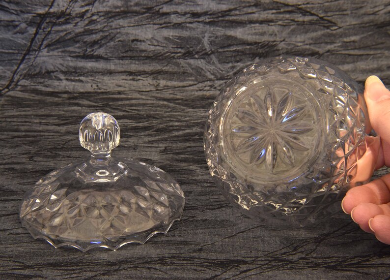 May include: A clear, cut-glass candy dish with a lid. The lid has a round, knob-like handle. The dish and lid feature a diamond pattern. The bottom of the dish has a starburst design.