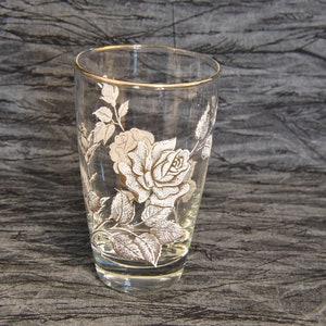 K&ouml;nnte beinhalten: Ein klares Glas Trinkglas mit goldfarbenem Rand und einem wei&szlig;en Blumendesign. Das Design zeigt eine Rose mit Bl&auml;ttern und Stielen.