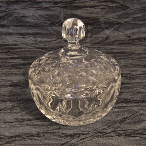 May include: A clear, round glass candy dish with a lid. The dish and lid are decorated with a diamond-shaped pattern. The lid has a small, round handle. The dish is sitting on a dark, textured surface.