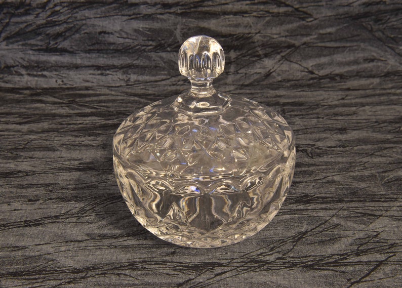 May include: A clear, round glass candy dish with a lid. The dish and lid are decorated with a diamond-shaped pattern. The lid has a small, round handle. The dish is sitting on a dark, textured surface.