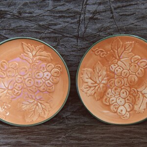 May include: Two small, orange ceramic plates with a green rim. Each plate features a raised design of a bunch of grapes with leaves.