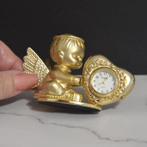 Puede incluir: Una figura dorada de querubín sosteniendo un reloj en forma de corazón. La esfera del reloj es blanca con números negros y manecillas doradas. El querubín tiene alas detalladas y está sentado.