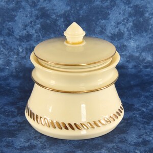 Puede incluir: Un tarro de cerámica blanco con tapa dorada y ribete dorado. El tarro tiene un diseño de cuerda alrededor de la parte inferior.