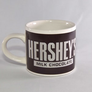 May include: A white ceramic mug with a brown band around the middle. The band has the words "HERSHEY'S" in white letters and "MILK CHOCOLATE" in smaller white letters.