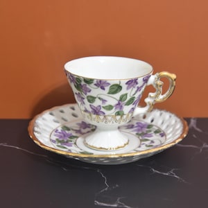 Puede incluir: Un juego de taza y platillo de té blanco con un diseño floral con violetas moradas y verdes. El platillo tiene un delicado patrón calado.
