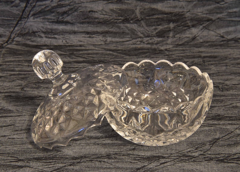 May include: A clear, cut-glass candy dish with a lid. The dish has a diamond pattern and a scalloped edge. The lid has a knob handle. The dish is open, revealing its interior. The overall theme is vintage glassware.