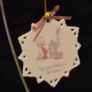Könnte beinhalten: Weißer Weihnachtsornament aus Keramik mit Schneeflockenmuster. Es zeigt eine Illustration eines Kindes und eines Ofens mit dem Text "May Your Christmas Be Warm". Der Ornament ist mit einer goldenen Glocke und einem rosa Band verziert.