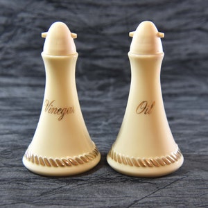 Puede incluir: Dos botellas de aceite y vinagre de cerámica blanca con ribetes dorados y tapas. Las botellas están etiquetadas como "Aceite" y "Vinagre".