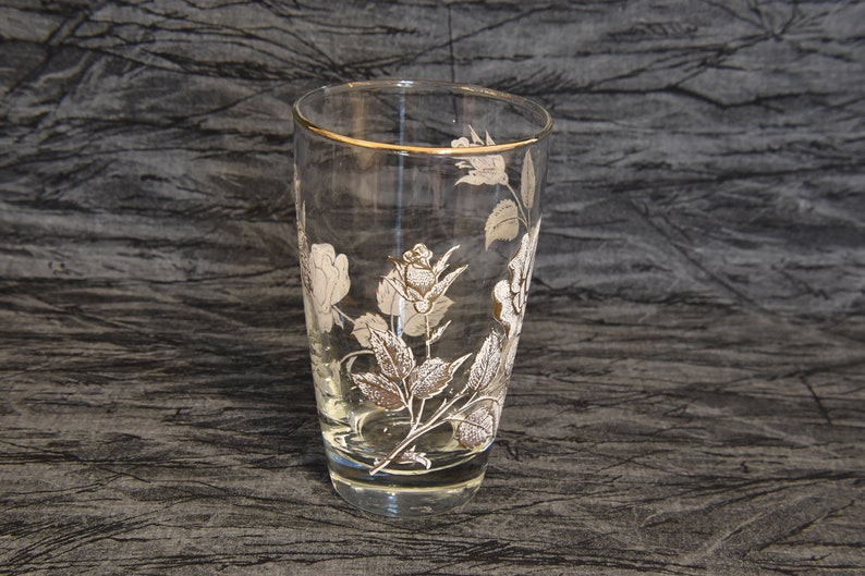 K&ouml;nnte beinhalten: Ein klares Glas mit goldfarbenem Rand und einem wei&szlig;en Blumenmuster. Die Blumen sind Rosen mit Bl&auml;ttern und Stielen.