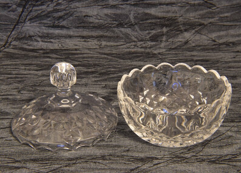 May include: A clear, cut-glass candy dish with a lid. The dish has a scalloped edge and a diamond pattern. The lid has a round knob. The dish is designed to hold small candies or other treats.
