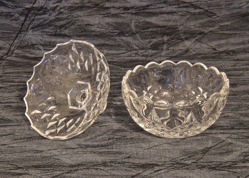 May include: Two clear, cut-glass bowls. One bowl is round with a scalloped edge, and the other is a lid with a hexagonal knob. Both have a diamond pattern. The bowls are likely for serving or display.