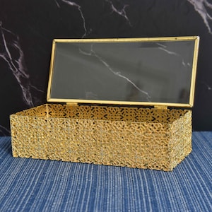 May include: A rectangular gold-colored decorative box with an open lid. The box has an intricate, openwork design on the sides and a clear top. The lid is framed in gold. The box is on a blue surface.