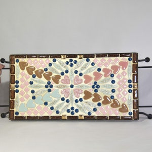 Puede incluir: Una bandeja de servir de mosaico decorativa con un marco marrón y un diseño de corazones rosas, marrones y azules sobre un fondo blanco. La bandeja está sobre un soporte de metal negro con dos asas.